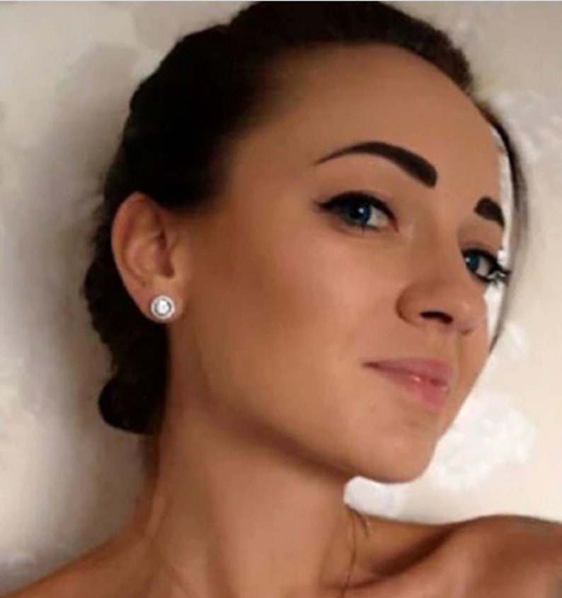 May include: A close-up shot of a person wearing a pair of round, silver stud earrings with a white, circular center stone and a halo of smaller stones. The earrings are set against the person's skin and dark hair.