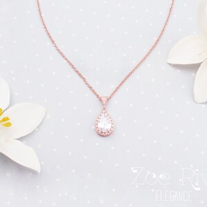 Peut inclure: Un collier en or rose avec un pendentif en forme de poire orn&eacute; de cristaux transparents et d'un halo de petits cristaux. Le collier est pr&eacute;sent&eacute; sur un fond bleu clair &agrave; pois blancs, avec des fleurs blanches de chaque c&ocirc;t&eacute;.