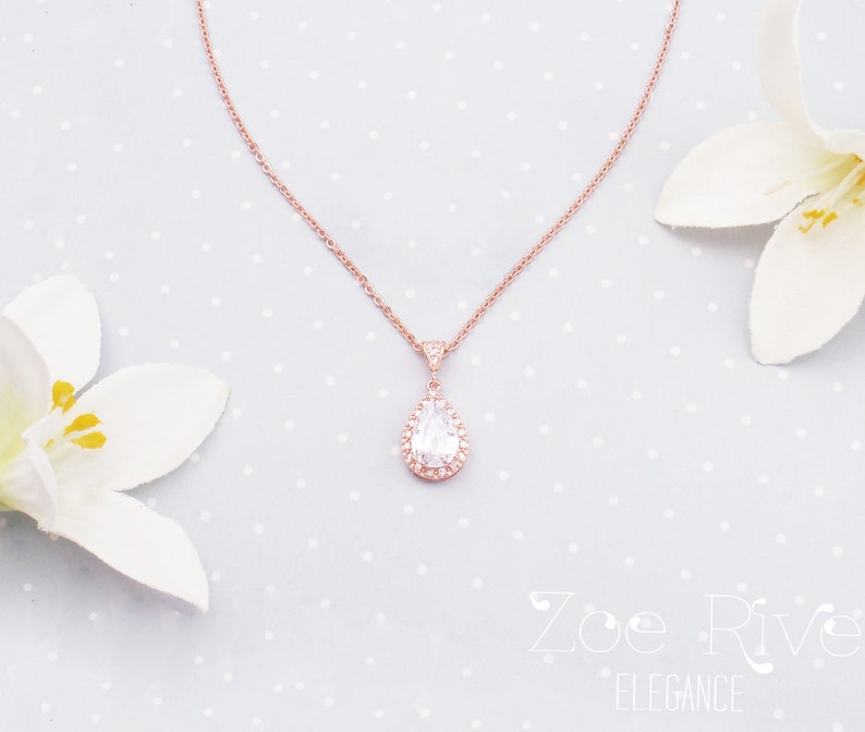 Peut inclure: Un collier en or rose avec un pendentif en forme de poire orn&eacute; de cristaux transparents et d'un halo de petits cristaux. Le collier est pr&eacute;sent&eacute; sur un fond bleu clair &agrave; pois blancs, avec des fleurs blanches de chaque c&ocirc;t&eacute;.