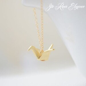 May include: A gold-colored origami crane pendant necklace. The pendant is suspended from a delicate chain. The necklace is displayed against a soft, white background. The text "Zoe River Elegance" is in the upper right corner.