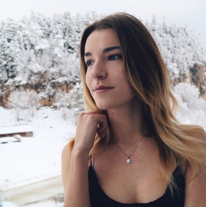 May include: A woman with blonde hair and a black top looks out at a snowy mountain landscape. She wears a silver necklace with a small, round pendant. The background features snow-covered trees and a cloudy sky.