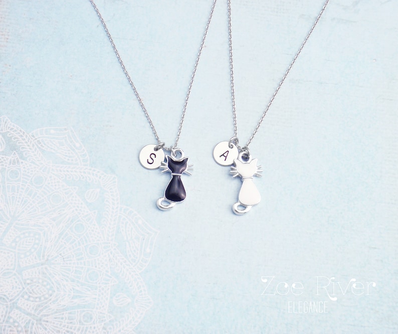 May include: Two silver cat pendant necklaces. One cat pendant is black, the other is white. Each necklace has a small circular charm with a letter. The background is light blue with a white floral design.