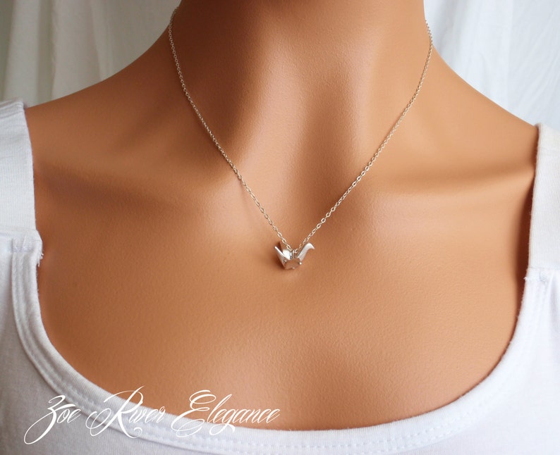 May include: A delicate silver necklace featuring a small, origami-style bird pendant. The pendant is suspended from a fine silver chain. The necklace is worn against a white top, with the brand name "Zoe River Elegance" in cursive script at the bottom.