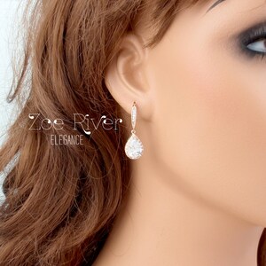 Peut inclure: Boucles d'oreilles pendantes en or rose avec un pendentif en cristal clair en forme de poire. Les boucles d'oreilles sont orn&eacute;es de petits cristaux &eacute;tincelants le long de la boucle et du haut du pendentif. Le texte "Zoe River Elegance" est visible.