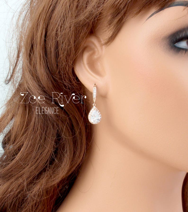 Peut inclure: Boucles d'oreilles pendantes en or rose avec un pendentif en cristal clair en forme de poire. Les boucles d'oreilles sont orn&eacute;es de petits cristaux &eacute;tincelants le long de la boucle et du haut du pendentif. Le texte "Zoe River Elegance" est visible.