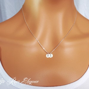 Op de afbeelding: Een fijne zilveren ketting met twee kleine, ronde hangers. Elke hanger heeft een enkele initiaal, "T" en "A". De ketting wordt gedragen om de hals van een persoon, tegen een witte top. De merknaam "Zoe River Elegance" staat onderaan.