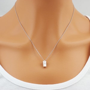 K&ouml;nnte beinhalten: Eine silberfarbene Halskette mit einem kleinen Anh&auml;nger in Form eines Kaffeebechers. Der Anh&auml;nger hat ein detailliertes Design und h&auml;ngt an einer feinen Kette. Die Halskette wird &uuml;ber einem wei&szlig;en Oberteil getragen.