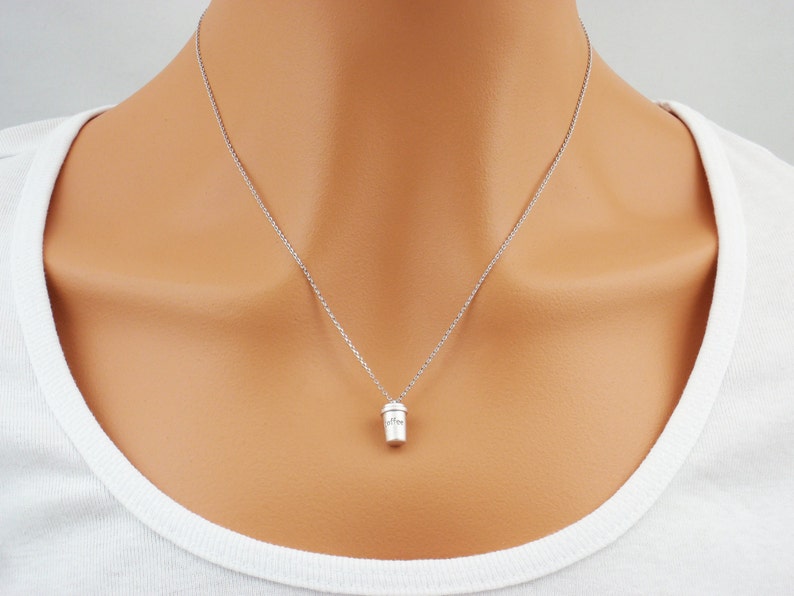 K&ouml;nnte beinhalten: Eine silberfarbene Halskette mit einem kleinen Anh&auml;nger in Form eines Kaffeebechers. Der Anh&auml;nger hat ein detailliertes Design und h&auml;ngt an einer feinen Kette. Die Halskette wird &uuml;ber einem wei&szlig;en Oberteil getragen.