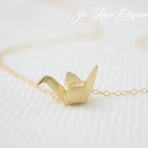 May include: A delicate gold-colored necklace featuring a small origami crane pendant. The crane is intricately detailed, with visible folds and a polished finish. The chain is fine and subtle, complementing the pendant's design. The necklace is set against a soft, white background.