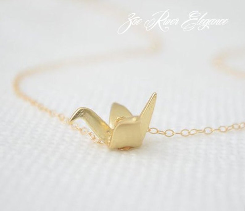 May include: A delicate gold-colored necklace featuring a small origami crane pendant. The crane is intricately detailed, with visible folds and a polished finish. The chain is fine and subtle, complementing the pendant's design. The necklace is set against a soft, white background.