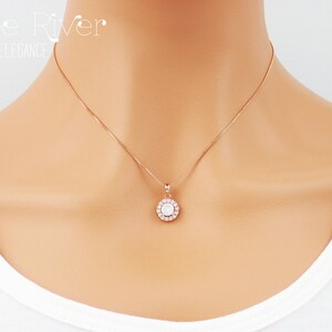 May include: Rose gold necklace with a round pendant featuring a central clear stone surrounded by smaller clear stones. The necklace is delicate and hangs on a thin chain. The text "Zoe River Elegance" is in the upper left corner.