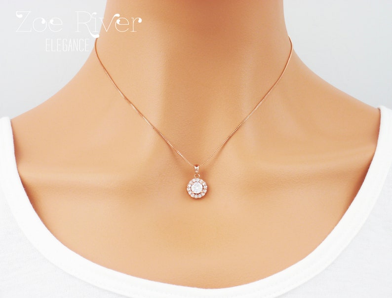 May include: Rose gold necklace with a round pendant featuring a central clear stone surrounded by smaller clear stones. The necklace is delicate and hangs on a thin chain. The text "Zoe River Elegance" is in the upper left corner.