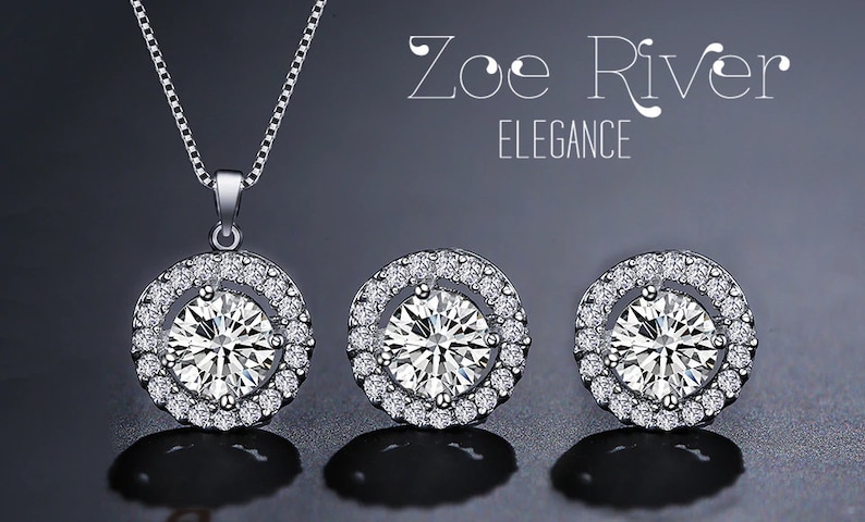 May include: A silver necklace and matching earrings set. The pendant and earrings feature a large, round, clear gemstone surrounded by a halo of smaller, sparkling stones. The set is displayed on a dark background, with the text "Zoe River ELEGANCE".