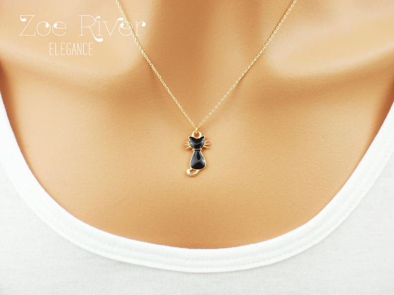 May include: A gold-colored necklace with a black cat pendant. The pendant is a stylized cat silhouette with a detailed face and tail. The necklace is displayed against a white background, with the text "Zoe River Elegance" in the upper left corner.