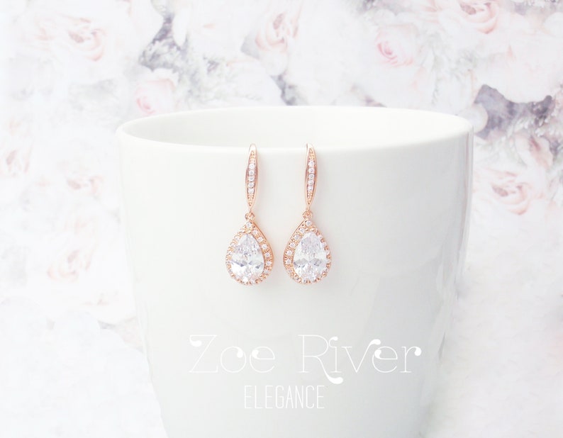 Peut inclure: Boucles d'oreilles en forme de goutte d'eau en or rose, avec un halo de petits cristaux transparents autour d'un grand cristal clair en forme de poire. Les boucles d'oreilles sont pr&eacute;sent&eacute;es sur une surface blanche. Le texte "Zoe River Elegance" est visible.