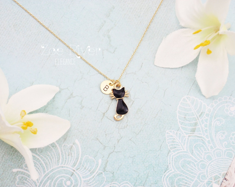 May include: A gold-colored necklace featuring a black cat pendant with a gold-colored collar and whiskers. The necklace also includes a small, round charm with the letter "B". The background is a light blue color with white floral accents.