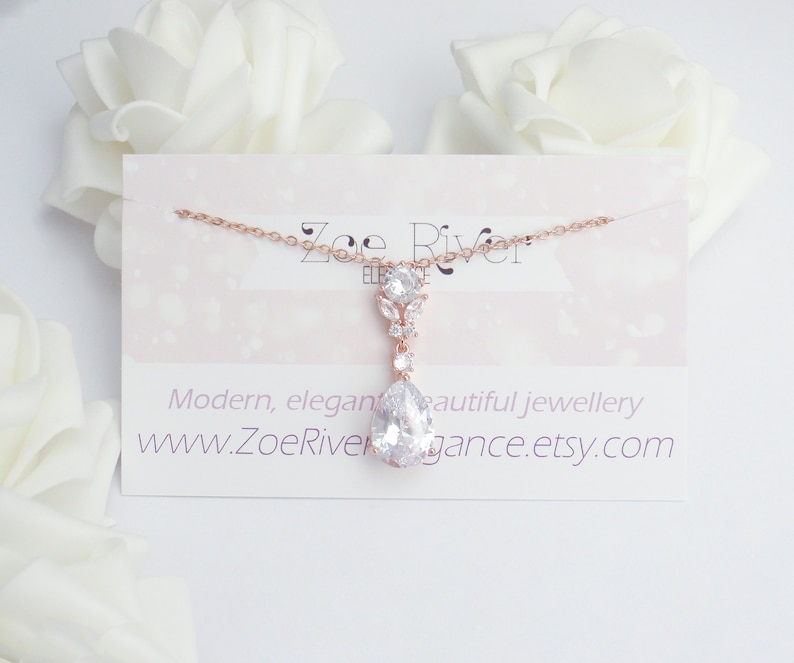 May include: A rose gold necklace with a large, pear-shaped clear crystal pendant. The pendant is suspended from a delicate chain and is accompanied by smaller, clear crystals. The necklace is displayed on a card with the text "Modern, elegant beautiful jewellery".