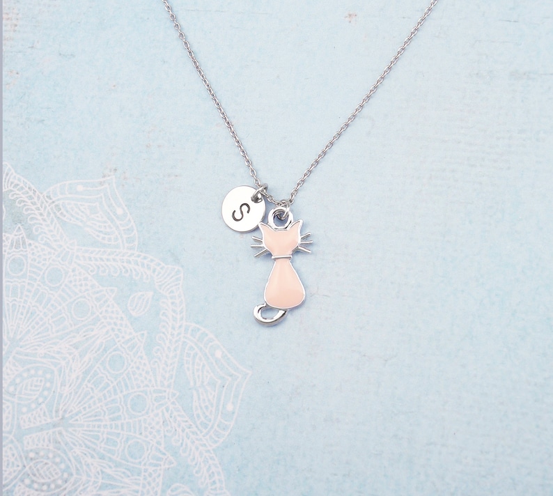 May include: A silver necklace featuring a light pink cat pendant and a circular charm with the letter "S". The cat pendant has a detailed design with a collar. The necklace is displayed on a light blue background.
