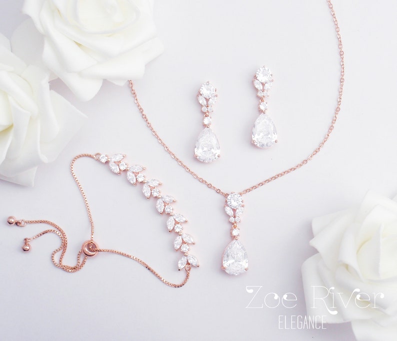 May include: A rose gold jewelry set featuring a bracelet, necklace, and earrings. The bracelet and earrings have a leaf design, while the necklace and earrings have a teardrop-shaped pendant. The jewelry is adorned with clear crystals.