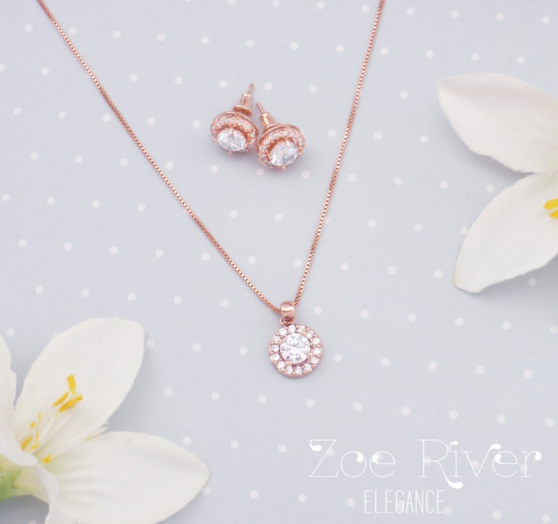 May include: A rose gold jewelry set featuring a necklace with a round pendant and matching stud earrings. Each piece has a central clear stone surrounded by smaller stones. The set is displayed on a light blue background with white polka dots.