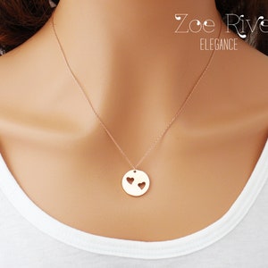May include: A gold-colored pendant necklace with a delicate chain. The pendant is a circular disc with two heart-shaped cutouts. The necklace is worn against a white top. The text "Zoe River Elegance" is visible.