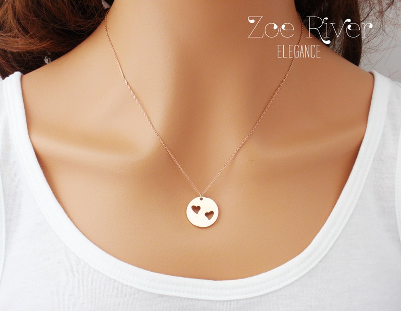 May include: A gold-colored pendant necklace with a delicate chain. The pendant is a circular disc with two heart-shaped cutouts. The necklace is worn against a white top. The text "Zoe River Elegance" is visible.