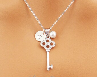 May include: A silver necklace with a key pendant, a pearl, and a circular charm with the letter "C". The key has a floral design and is embellished with small stones. The necklace is suspended from a fine chain.