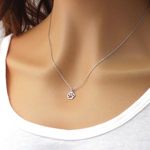May include: A silver necklace with a small paw print pendant. The pendant, a detailed cutout of a paw, hangs from a fine chain. The necklace is displayed against a white background.