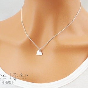May include: A delicate silver necklace featuring a small heart pendant. The heart-shaped charm hangs from a fine chain, creating a minimalist and elegant look. The necklace is worn against a white top.