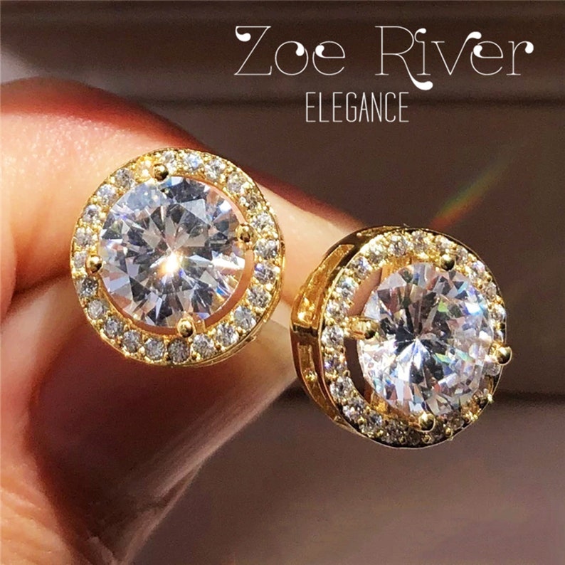 May include: A pair of gold-colored stud earrings with a large, clear, round center stone surrounded by smaller, clear stones. The earrings are set in a gold-colored metal and the text "Zoe River ELEGANCE" is visible.