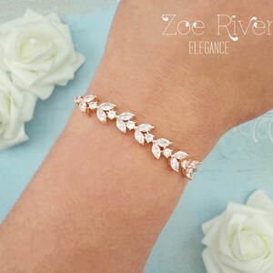 May include: A gold-toned bracelet with a leaf design, adorned with clear, sparkling crystals. The bracelet is displayed on a wrist, with the text "Zoe River Elegance" visible in the background. White roses are also in the background.