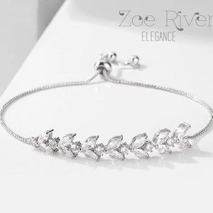 May include: A silver bracelet with a delicate leaf design. The bracelet features clear, sparkling gemstones set in a row. The text "Zoe River Elegance" is displayed in the upper portion of the image.