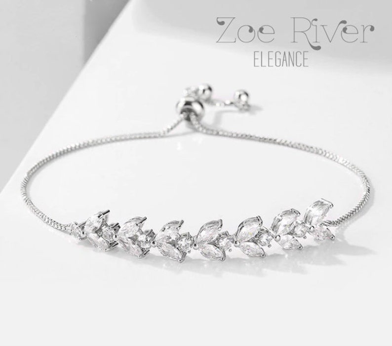 May include: A silver bracelet with a delicate leaf design. The bracelet features clear, sparkling gemstones set in a row. The text "Zoe River Elegance" is displayed in the upper portion of the image.