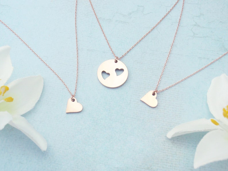 May include: Rose gold necklaces with heart pendants. One necklace features a round pendant with two heart cutouts. Two other necklaces have solid heart pendants. The necklaces are displayed on a light blue surface with white flowers.