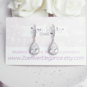 Peut inclure: Une paire de boucles d'oreilles pendantes argent&eacute;es avec un pendentif en cristal clair en forme de poire. Les boucles d'oreilles sont pr&eacute;sent&eacute;es sur une carte avec le texte "Zoe River Elegance" et une adresse de site web. Des fleurs blanches sont en arri&egrave;re-plan.