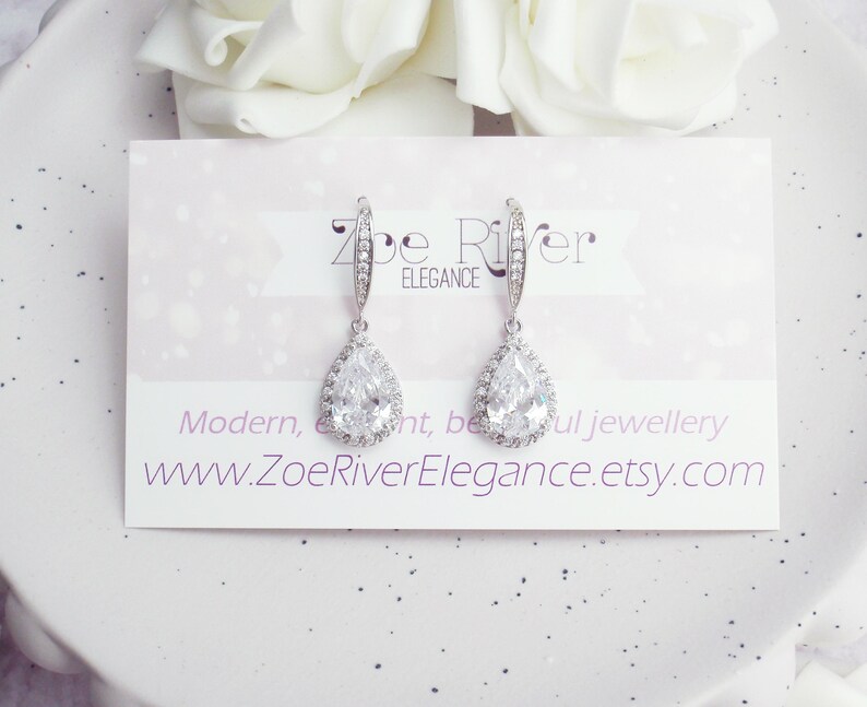 Peut inclure: Une paire de boucles d'oreilles pendantes argent&eacute;es avec un pendentif en cristal clair en forme de poire. Les boucles d'oreilles sont pr&eacute;sent&eacute;es sur une carte avec le texte "Zoe River Elegance" et une adresse de site web. Des fleurs blanches sont en arri&egrave;re-plan.