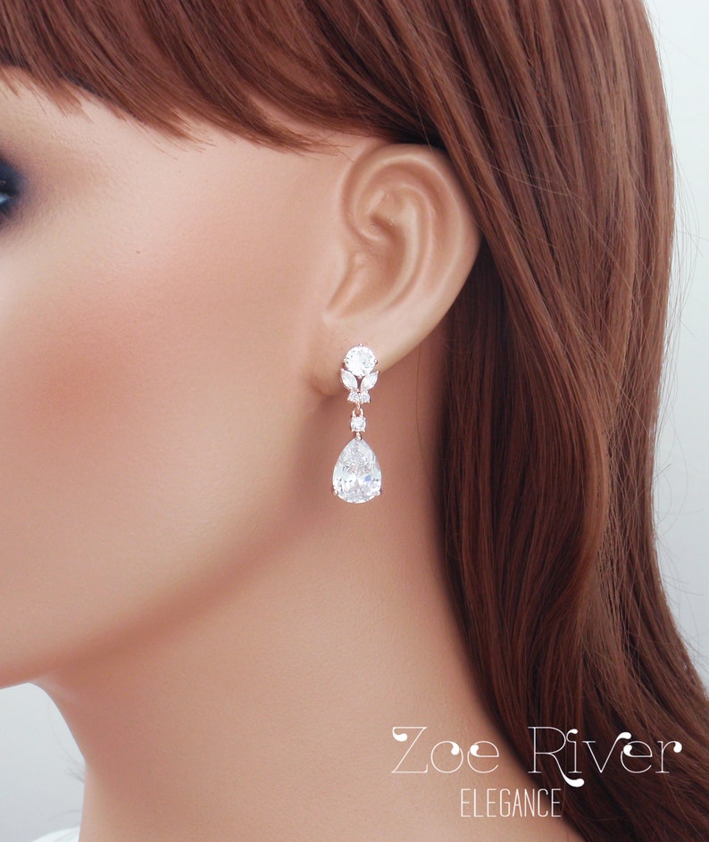 May include: Elegant silver-toned drop earrings with sparkling clear crystals. The earrings feature a round stud, a floral design, and a large pear-shaped crystal. The earrings are from the Zoe River Elegance collection.