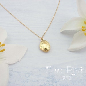 May include: A gold-colored oval locket necklace with a delicate chain. The locket has an ornate design and is displayed with white flowers. The text "Zoe River Elegance" is visible in the image.