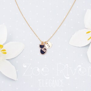 May include: Gold-tone necklace featuring a black cat pendant with a gold outline and a small round charm with the letter "S". The necklace is displayed on a light blue background with white flowers.