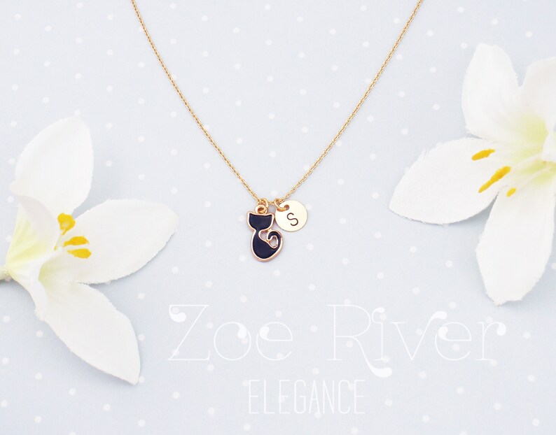 May include: Gold-tone necklace featuring a black cat pendant with a gold outline and a small round charm with the letter "S". The necklace is displayed on a light blue background with white flowers.