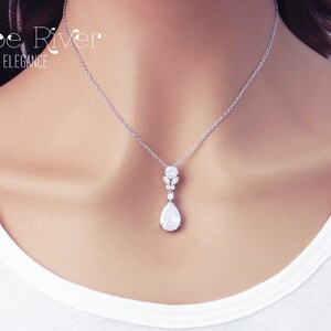 May include: A silver pendant necklace with a pear-shaped, clear crystal drop. The necklace features a delicate chain and a cluster of smaller crystals above the drop. The text "Zoe River Elegance" is in the upper left corner.
