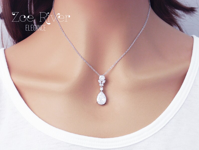 May include: A silver pendant necklace with a pear-shaped, clear crystal drop. The necklace features a delicate chain and a cluster of smaller crystals above the drop. The text "Zoe River Elegance" is in the upper left corner.