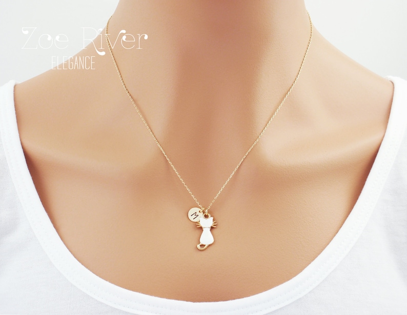 May include: A gold-colored necklace featuring a cat pendant with a small circular charm. The cat pendant is white with gold-colored details. The necklace is worn against a white shirt. The text "Zoe River Elegance" is in the upper left corner.