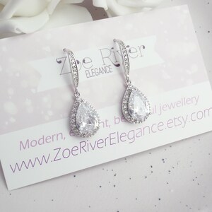 Peut inclure: Paire de boucles d'oreilles pendantes argent&eacute;es avec une pierre centrale en cristal clair en forme de poire, entour&eacute;e de petits cristaux clairs. Le texte "Zoe River Elegance" et l'adresse du site web sont visibles.