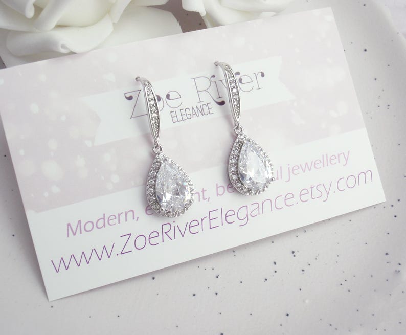 Peut inclure: Paire de boucles d'oreilles pendantes argent&eacute;es avec une pierre centrale en cristal clair en forme de poire, entour&eacute;e de petits cristaux clairs. Le texte "Zoe River Elegance" et l'adresse du site web sont visibles.