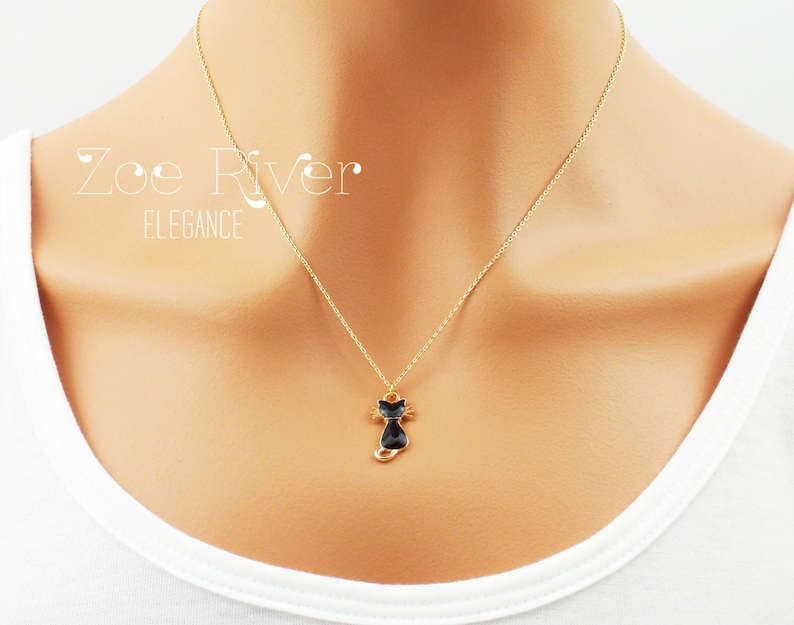May include: A gold-colored necklace with a small black cat pendant. The cat pendant has a gold outline and is suspended from a delicate chain. The text "Zoe River Elegance" is visible in the upper left corner.