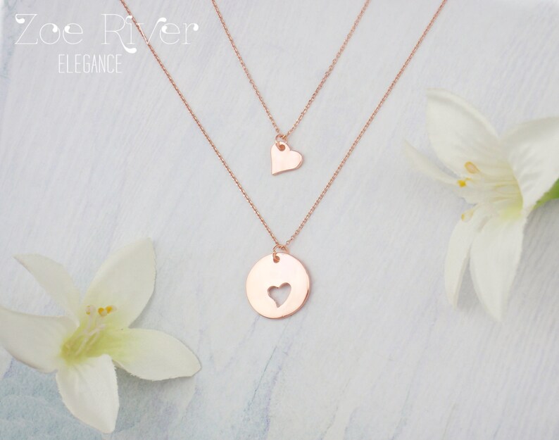 May include: Rose gold layered necklaces. The top necklace features a small heart pendant, while the lower necklace has a circular pendant with a heart cutout. The image includes the text "Zoe River Elegance" in the upper left corner. White flowers are in the background.
