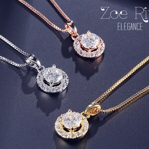 May include: Three necklaces with pendant designs. One is silver, one is rose gold, and one is gold. Each pendant features a large, clear, round gemstone surrounded by smaller stones. The necklaces are displayed on a dark, reflective surface. The text "Zoe River ELEGANCE" is in the upper right corner.