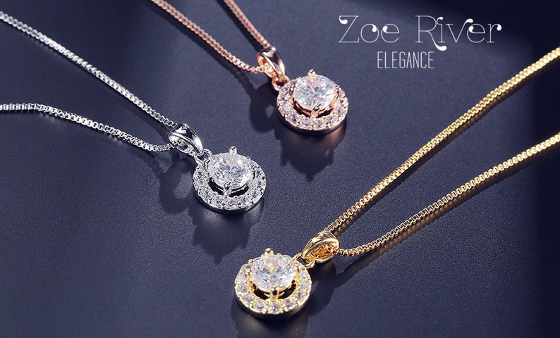 May include: Three necklaces with pendant designs. One is silver, one is rose gold, and one is gold. Each pendant features a large, clear, round gemstone surrounded by smaller stones. The necklaces are displayed on a dark, reflective surface. The text "Zoe River ELEGANCE" is in the upper right corner.