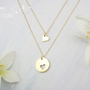 May include: Two gold-colored necklaces. The top necklace has a small heart pendant. The bottom necklace has a circular pendant with a heart-shaped cutout. The necklaces are layered and displayed with white flowers. The text "Zoe River Elegance" is in the upper left corner.
